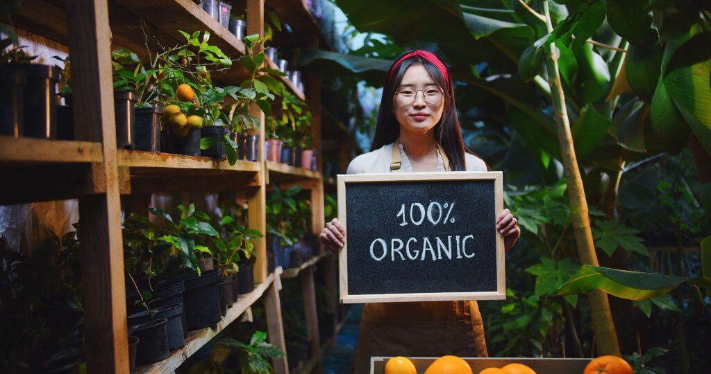 femme asiatique avec un bandeau rouge et des lunette vêtue d'un t-shirt blanc et dans tablier de travail orangé tient un tableau noir où est écrit : "100% Organic". Elle se trouve dans une serre ou pousse des arbres et des plantes, il y a aussi des agrumes qui poussent. 