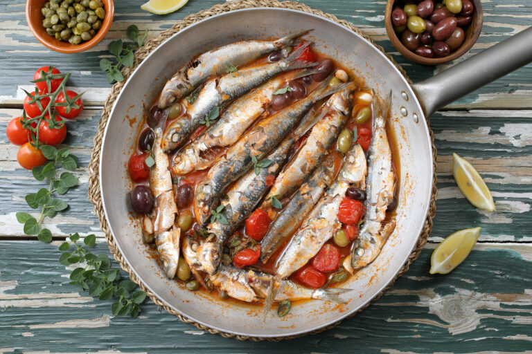 Plat cuisiné à base de sardines de tomates grillés, d’olives typique de la cuisine méditerranéenne. On trouve ce plat dans une poêle en inox au milieu de l’image, sur une planche de bois bleu en train de s’écailler. Autour du plat, on retrouve des quartiers de citron avec des herbes, des tomates cerise, des olives et des capres. 