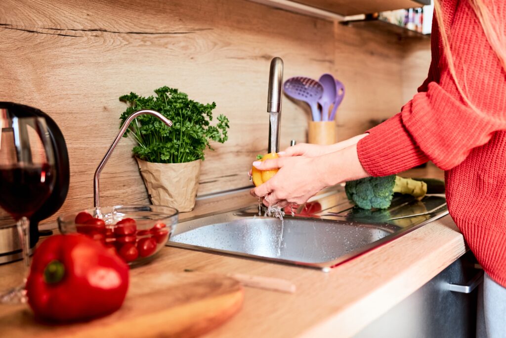 Femme avec un pull en laine rouge à gauche de l'image se trouve dans sa cuisine et lave un poivron de couleur jaune à l'au du robinet. la crédence de la cuisine est en bois ainsi que le plan de travail. L'évier est en inox. 