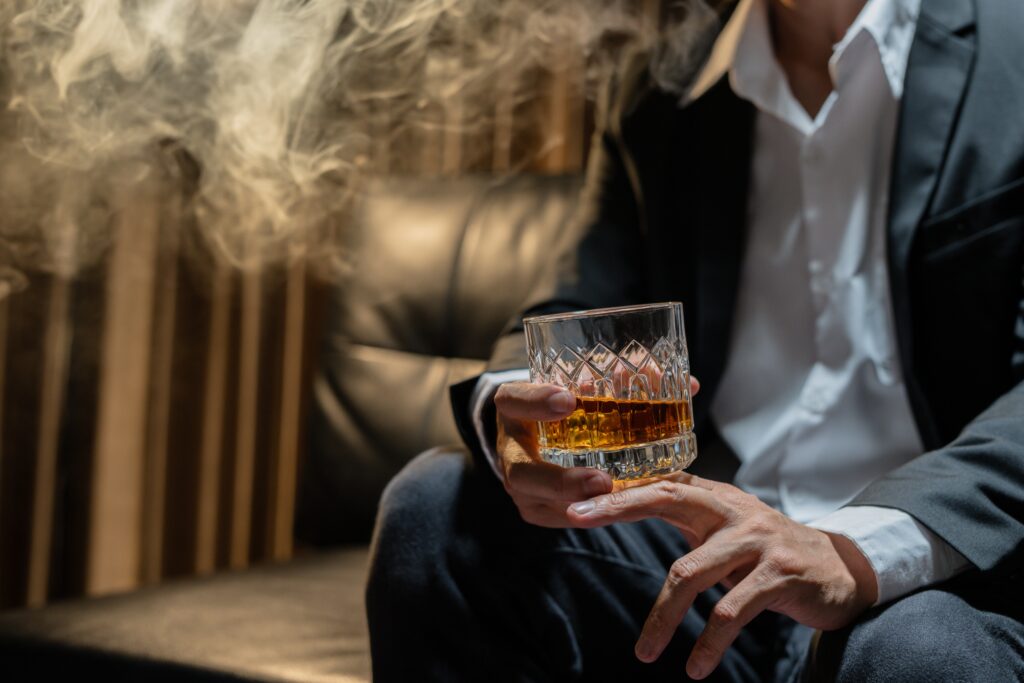 Un homme tient d'une main un verre de whisky, il y a de la fumée de cigarette. 