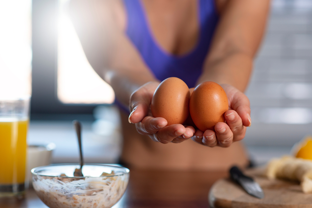 Au premier plan, un bol de porridge avec du fromage blanc à gauche de l’image. Au centre de l’image, les mains d’une jeune femme sportive tenant 2 œufs. 