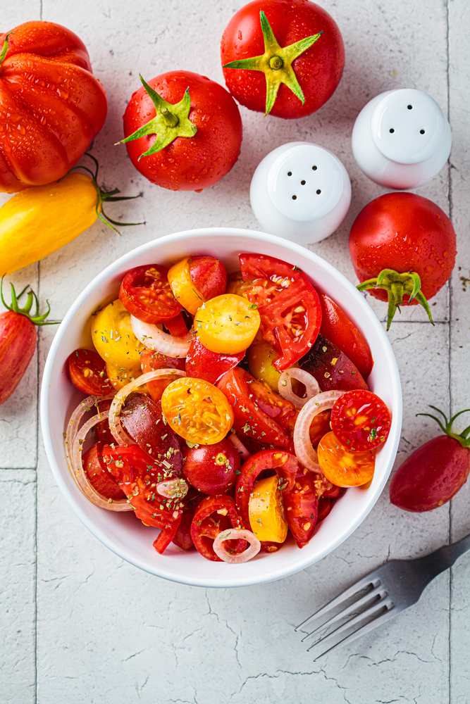 Salade de tomates et d’oignons avec des herbes aromatiques dans un bol blanc sur un sol blanc. La salade est rouge avec des nuances de jaune et des pointes de couleur verte. Les tomates sont coupées dans le bol et autour de ce bol sont posées des tomates entières de couleurs jaunes à rouges. Une salière, une poivrière ainsi qu’une fourchette complètent l’image.