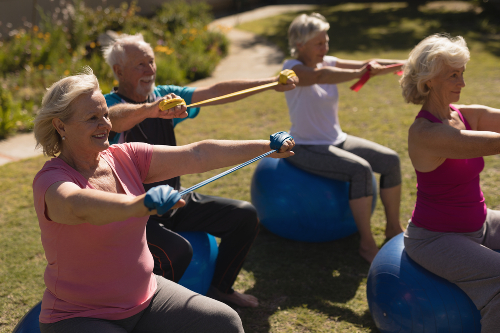 3 femmes âgées et un homme âgé en tenue de sport assis sur des ballons de gymnastique bleus effectuent des mouvements de résistance pour les épaules avec des élastiques.  