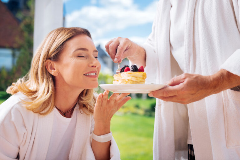 Femme âgée souriante à l'approche d'une assiette de pancakes servi par un homme en blanc. 