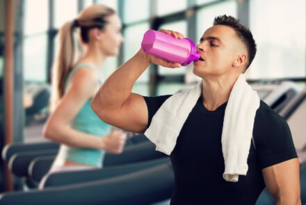 Au premier plan, un jeune homme en T-shirt noir et portant une serveiette autours du cou consomme une boisson dans une gourde rose. Il se trouve dans une salle de sport, en arrière plan une jeune femme cours sur un tapis.