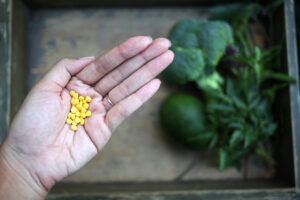 Une main de femme contient des comprimés d'acide folique. Des légumes verts sont posés sur une table en arrière plan.