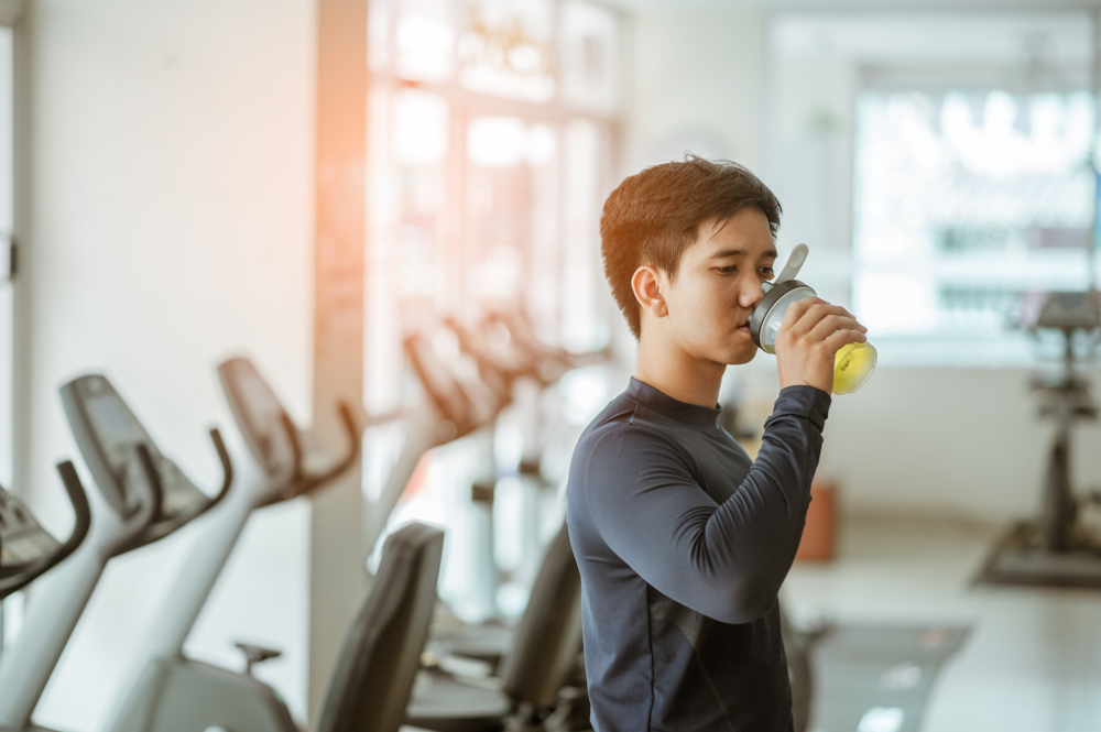 Jeune homme asiatique vêtu d'une tenue de sport grise est en train de boire un shaker de protéines après sa séance d'entrainement dans une salle de sport. 