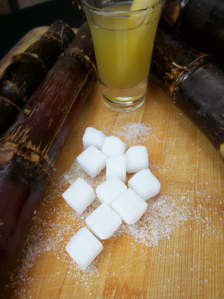 Sucre blanc issue de la canne à sucre sur une table en bois. Dans la partie supérieur, un verre contenant du sirop de sucre de canne, encadré de haut en bas et de droite à gauche par des morceau de tige brune de canne à sucre.